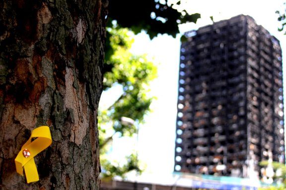 A tree with a yellow ribbon pinned to it, a burned block of flats in the background.